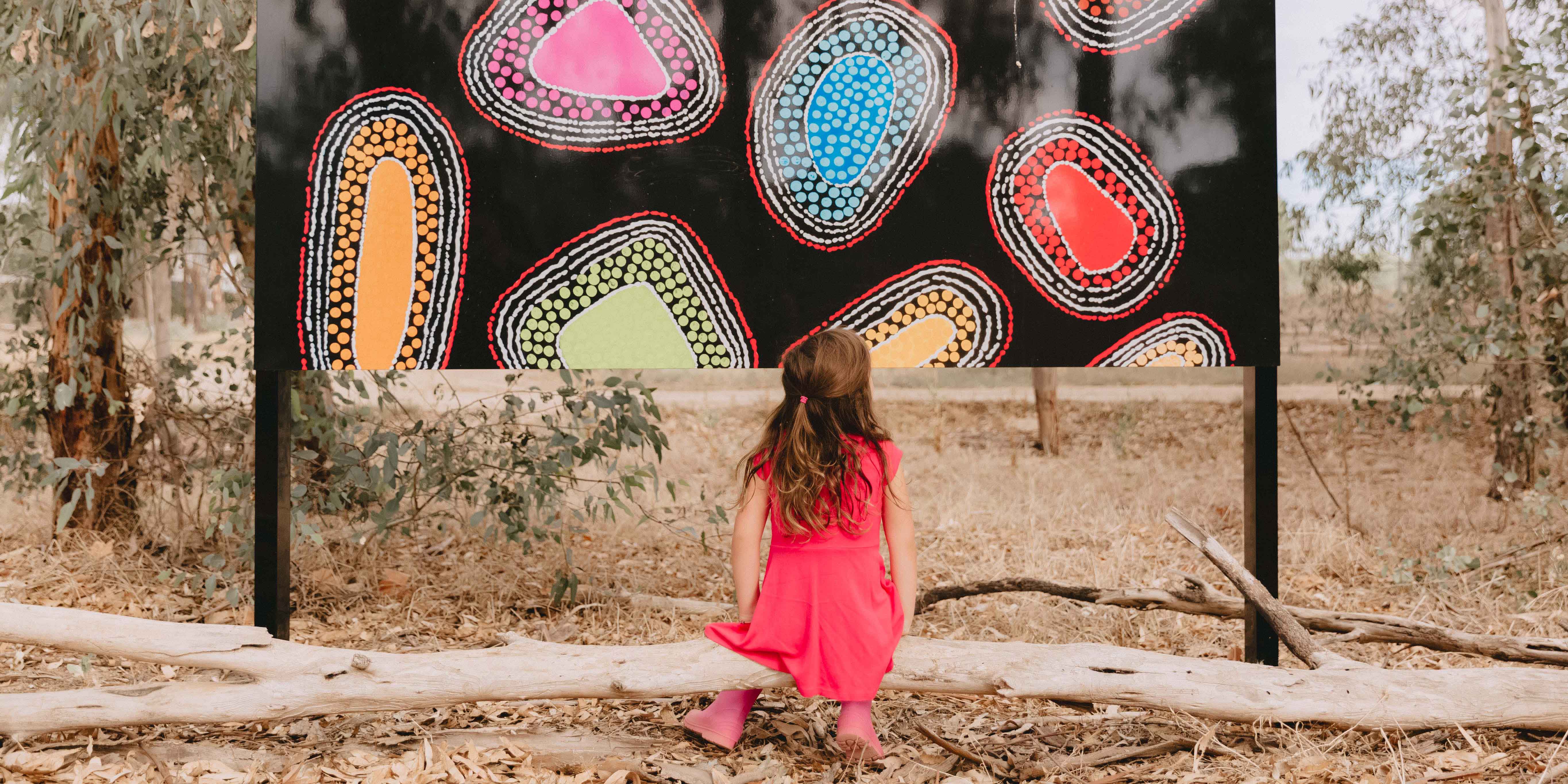 Girl sitting on a log looking up at a Wiradjuri painting at the entrance to Marrambidya Wetland in Wagga Wagga 