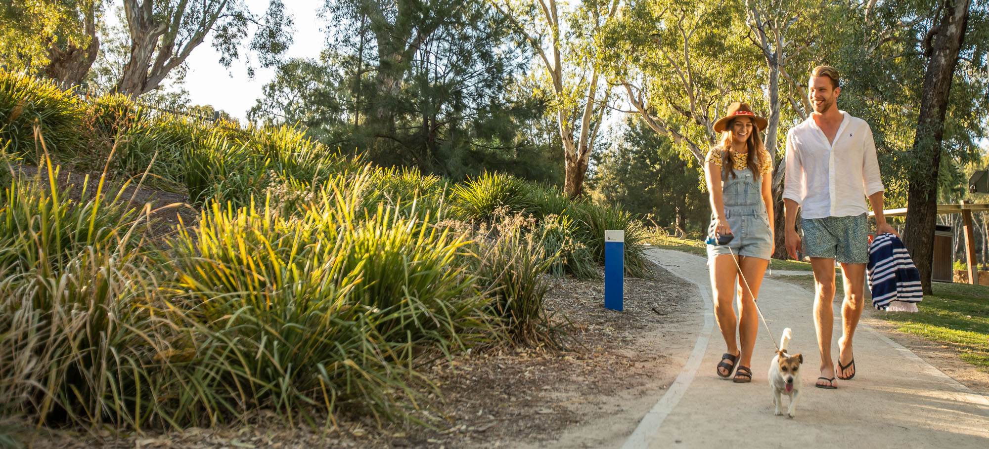 Female and male walking a small dog on a lead past Wagga Beach on the Wiradjuri Walking Track