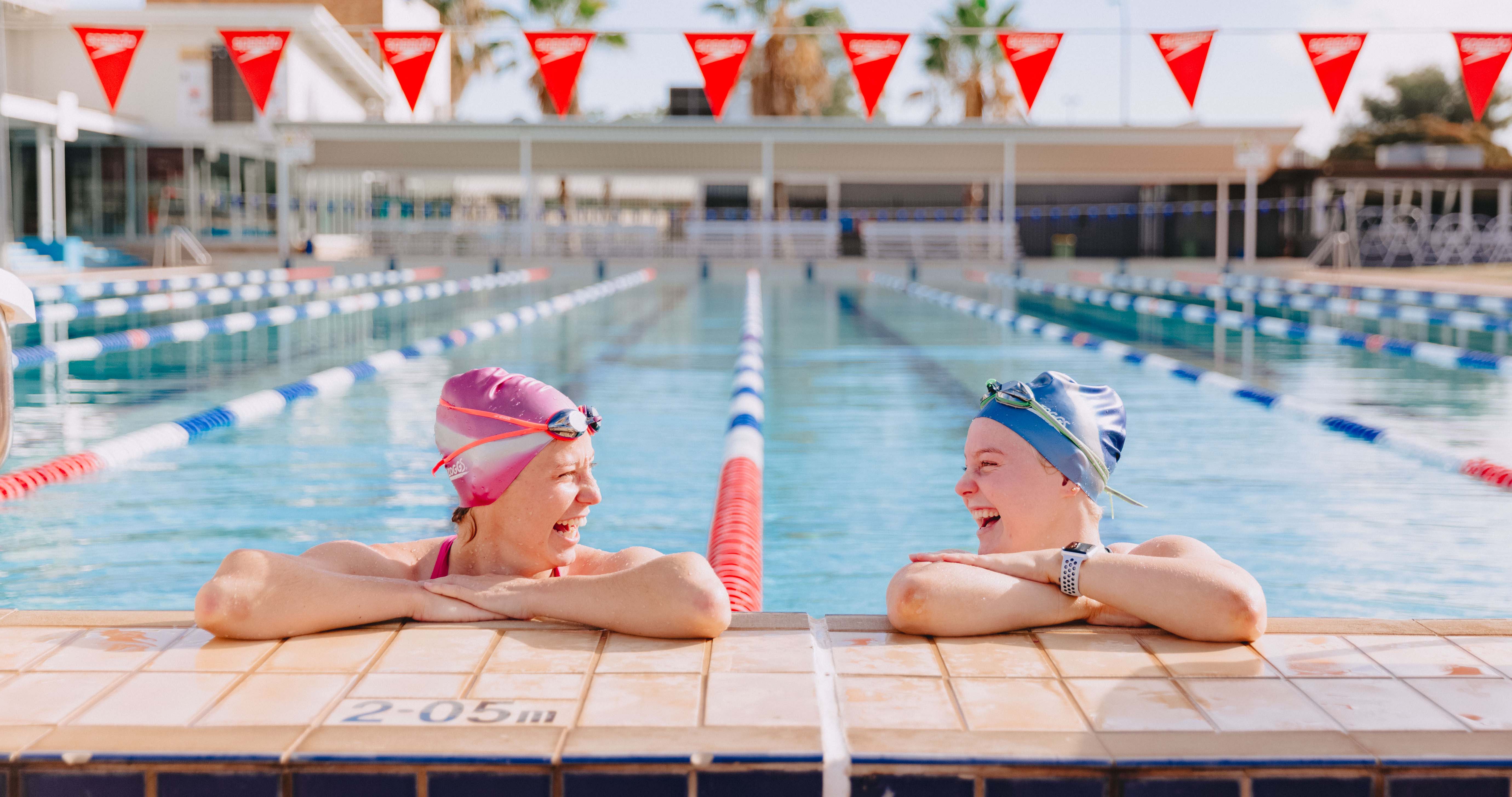 Two females leaning on the edge of the pool, laughing at each other. 